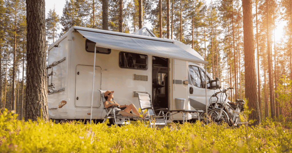 Woman relaxing in front of a parked motorhome in a forest, surrounded by tall trees and natural greenery.