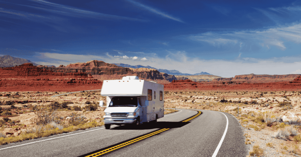 RV driving along a winding road through striking red rock formations in the American Southwest under a clear blue sky.
