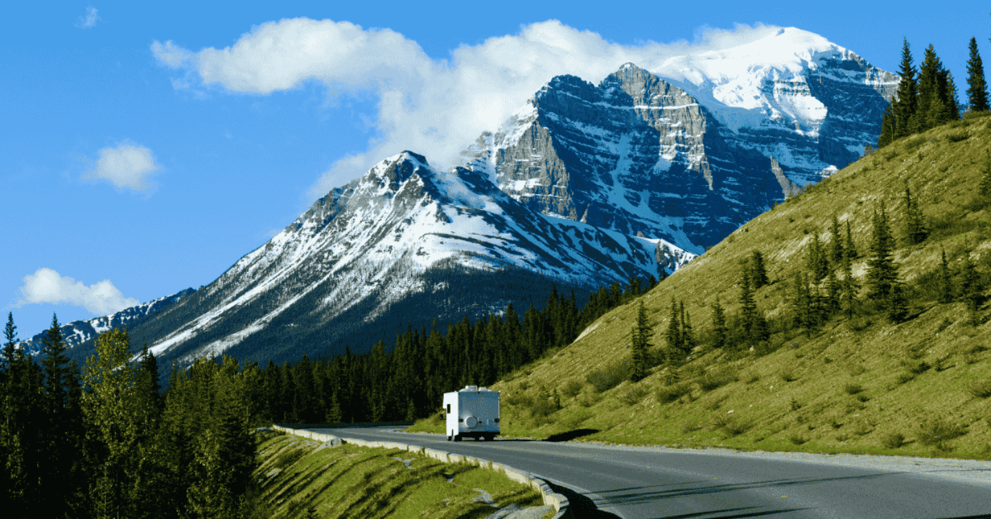 RV travelling along a scenic road in Canada with snow-covered mountains towering in the background under a clear sky.