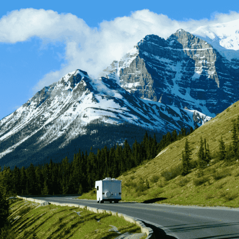 RV travelling along a scenic road in Canada with snow-covered mountains towering in the background under a clear sky.