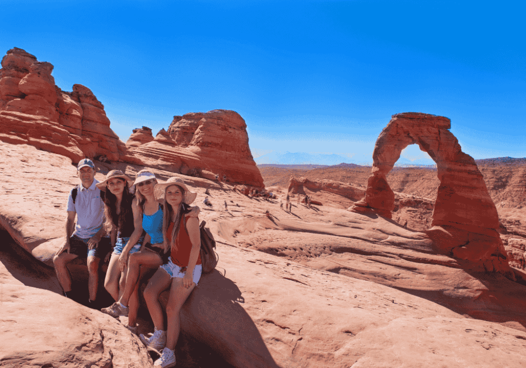A smiling family of four posing on a scenic trail surrounded by red rock formations and desert landscape in a Utah national park, enjoying a sunny day outdoors.
