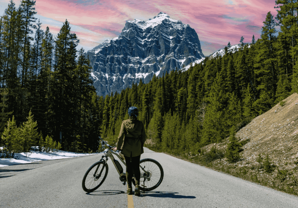 Cyclist standing on a mountain road with a bike, gazing at snow-covered peaks under a colorful sky.