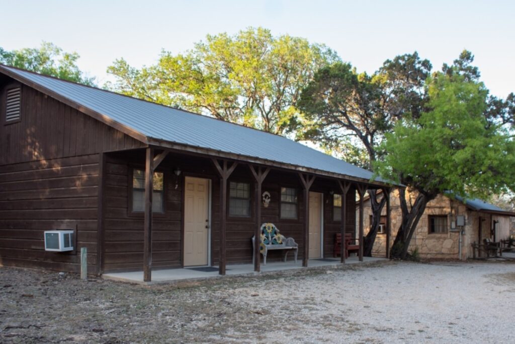 Exterior view of a rustic lodge at Mayan Dude Ranch in Bandera, Texas, surrounded by oak and cedar trees in scenic Hill Country